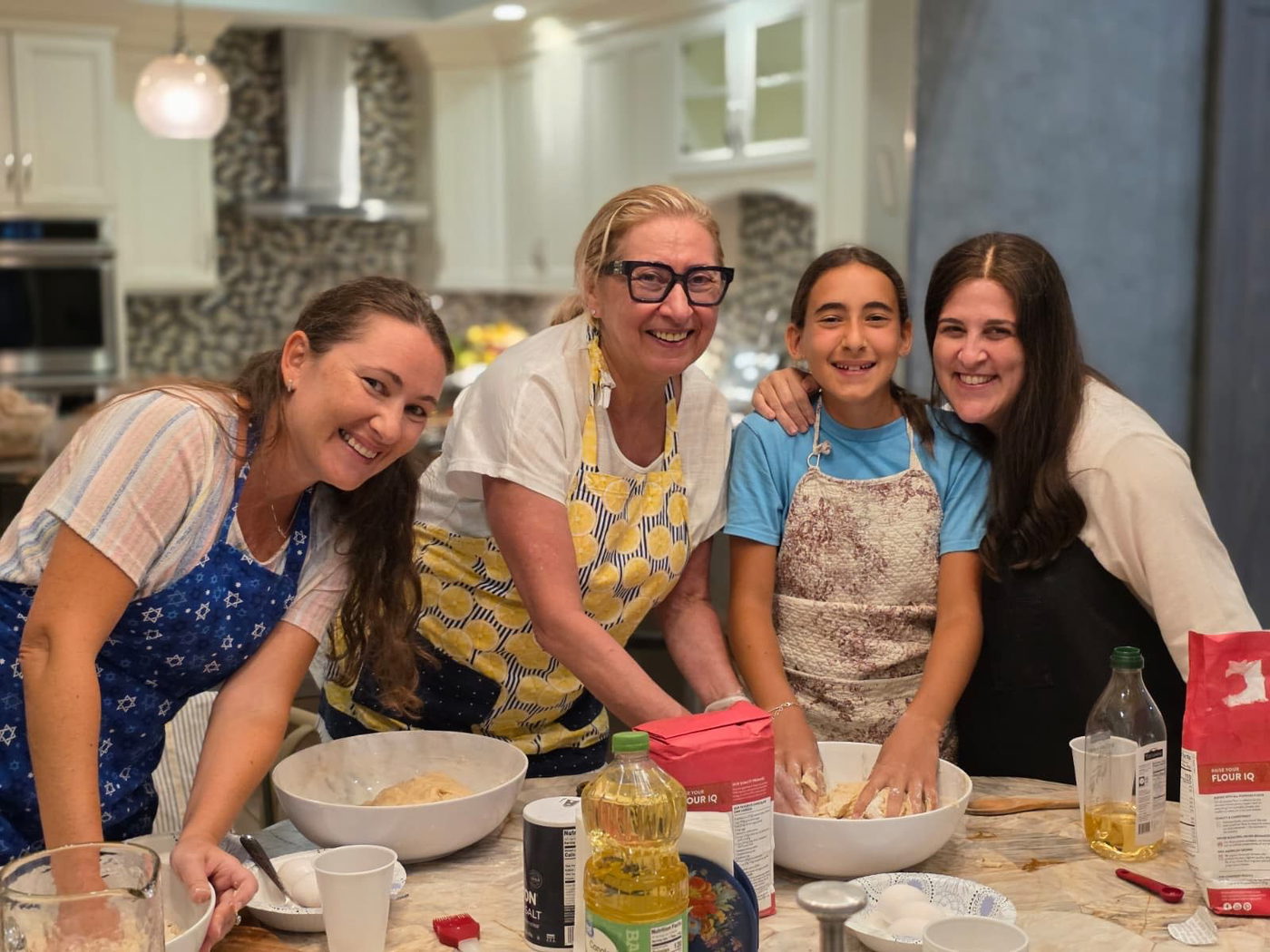 Women and a young girl in aprons mixing challah dough around a kitchen island