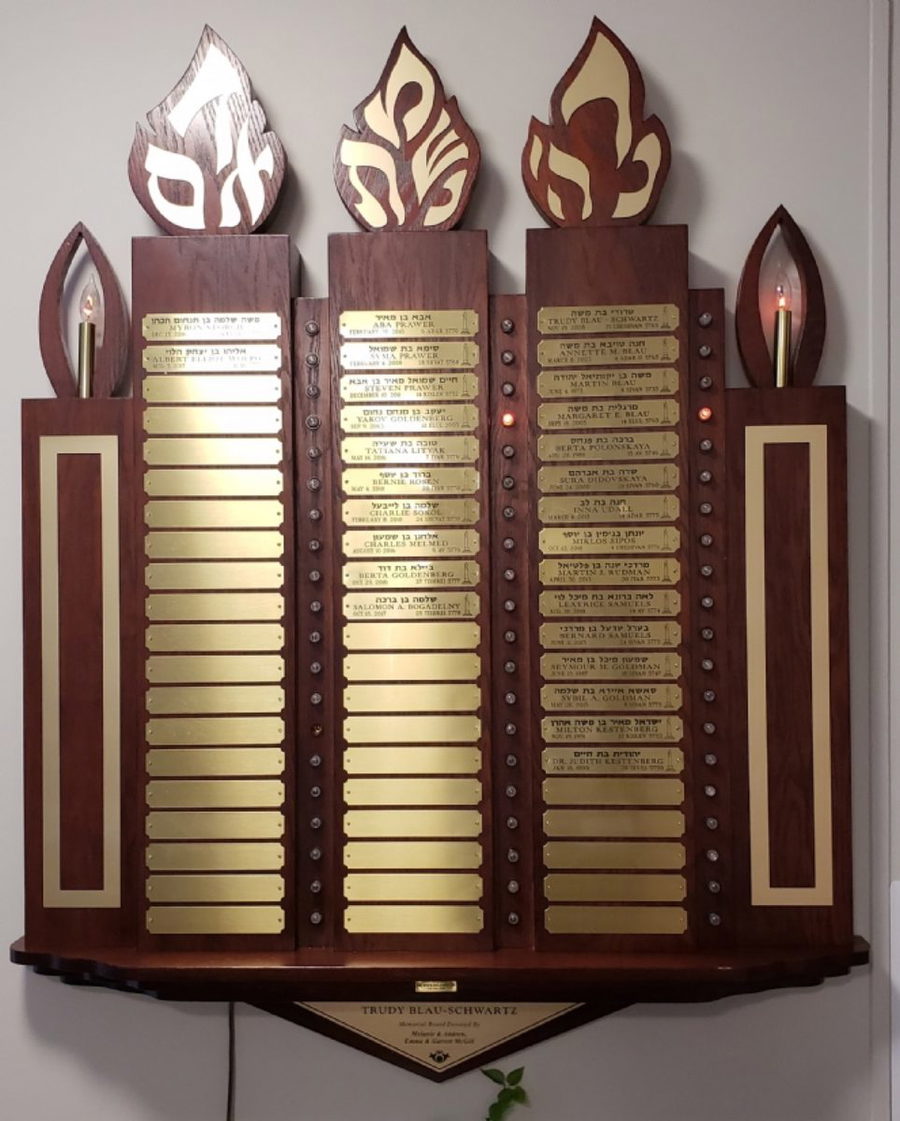 The memorial plaque wall at the Chabad House — carved wooden flames above columns of engraved name plates, each with a small memorial light