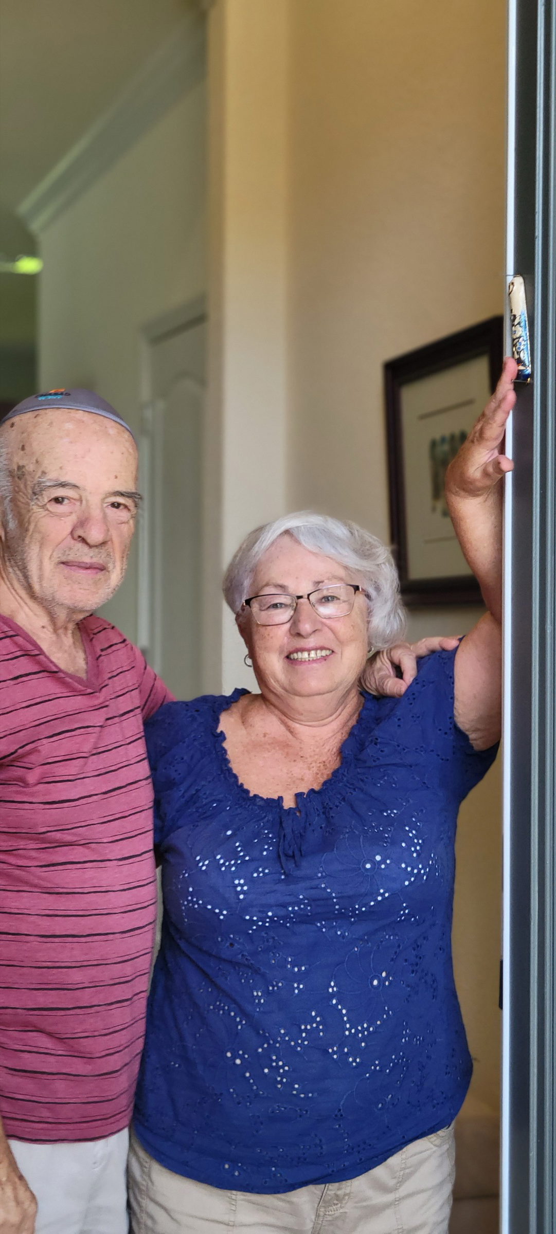 A smiling older couple standing together in their doorway beside their mezuzah